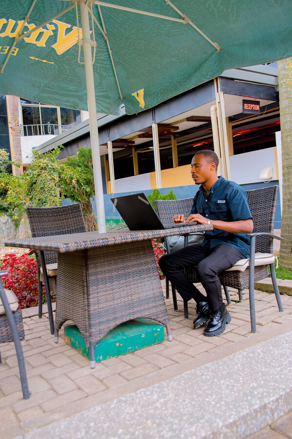 A man sitting at an outdoor table with a laptop, under a green umbrella, in front of a building with a reception sign. - INEZA HOTEL (Ruhengeri)