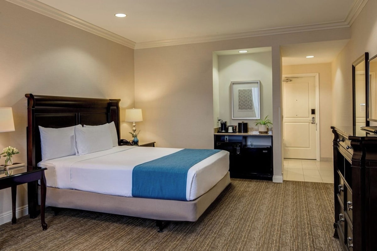 Habitación de hotel con una cama grande, mesitas de noche, un escritorio, un espejo y una puerta que da a otra habitación.