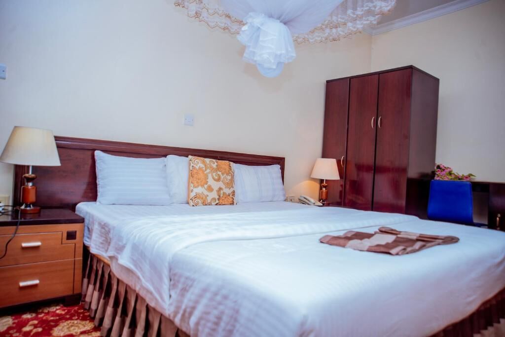 A neatly made bed with white linens and a patterned pillow, a wooden nightstand with a lamp, and a dark wooden wardrobe.