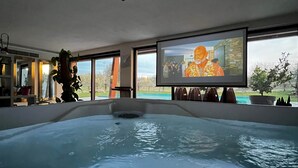 A hot tub with a projection screen displaying a person in traditional attire, surrounded by large windows and indoor plants. - Torre Menerva Viterbo Stunning Tuscia Villa,Private Spa + Sauna + Saltwater Pool (Lazio)