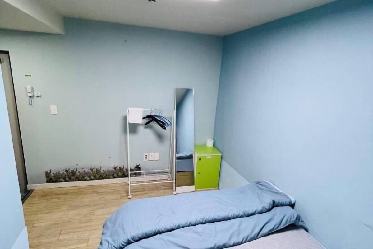 A room with a bed, a small green cabinet, a white shelf with a mirror, and a wall-mounted telephone.