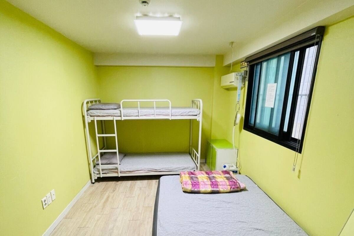 A small room with a bunk bed, a single bed, a window with curtains, and a ceiling light.