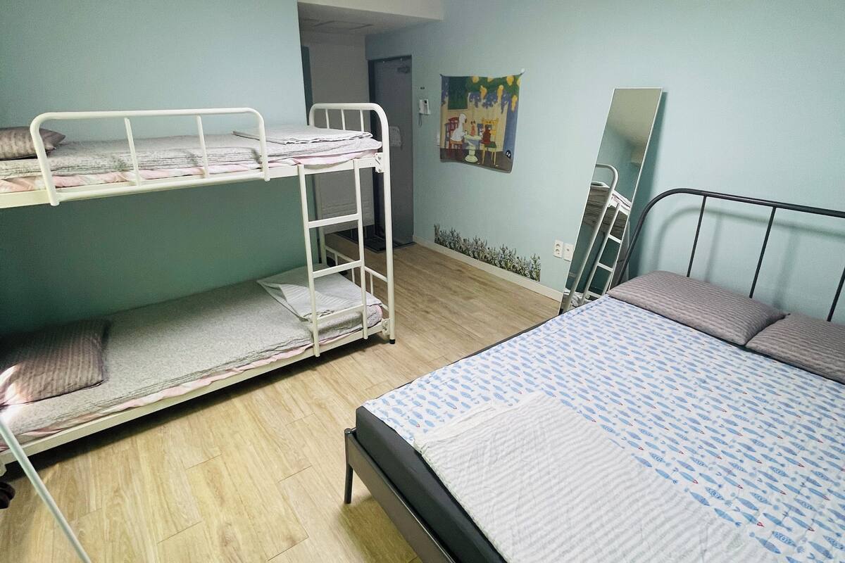A dormitory room with bunk beds, a bed, a mirror, and a door leading to another room.