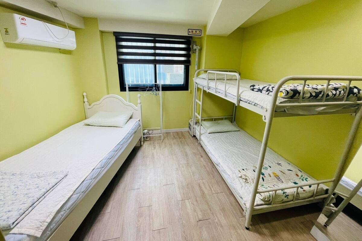 A dormitory room with bunk beds, a single bed, a window with blinds, an air conditioning unit, and a radiator.