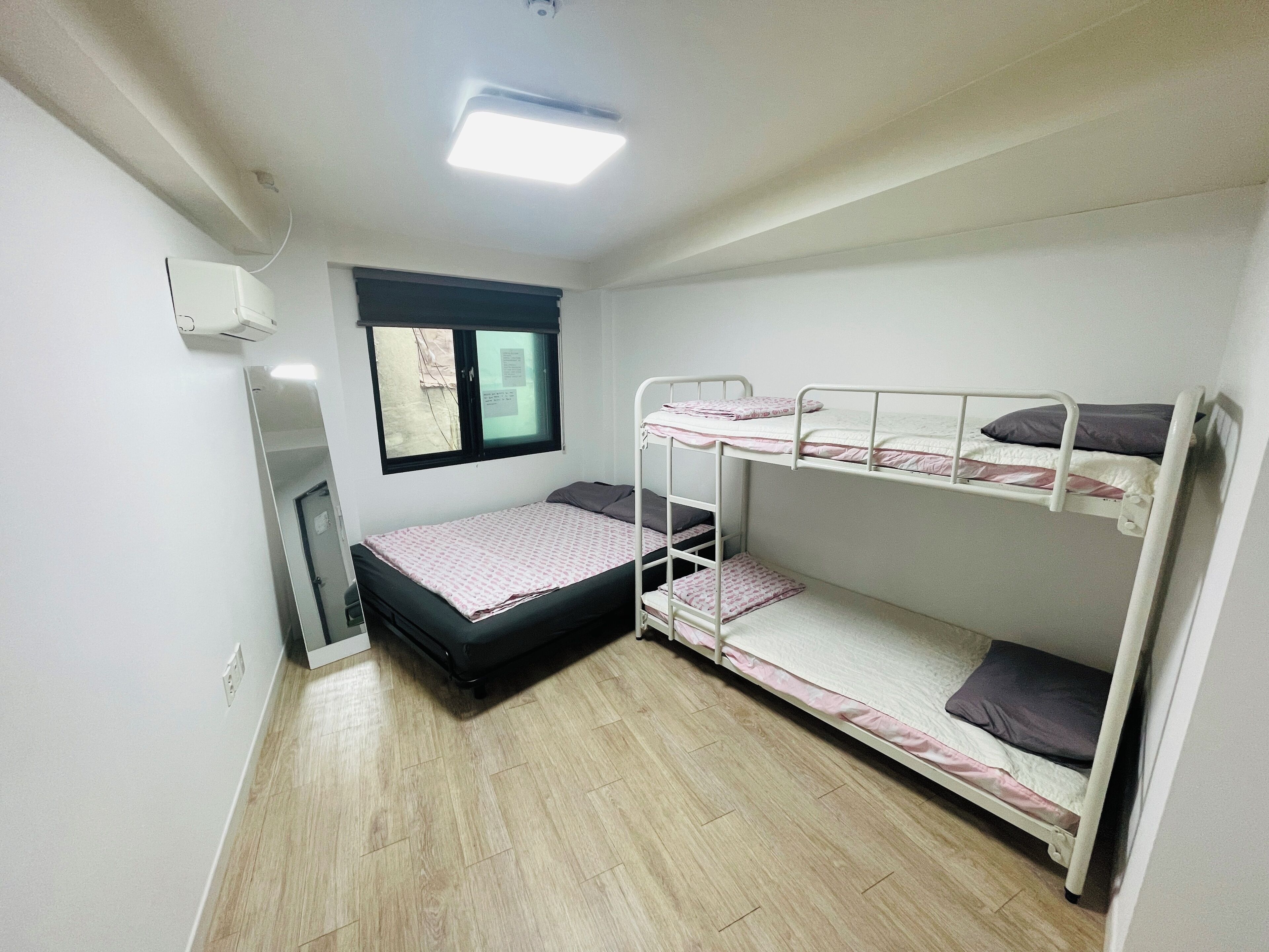 A small, clean room with a bunk bed, a single bed, a window with a view, and a ceiling light.