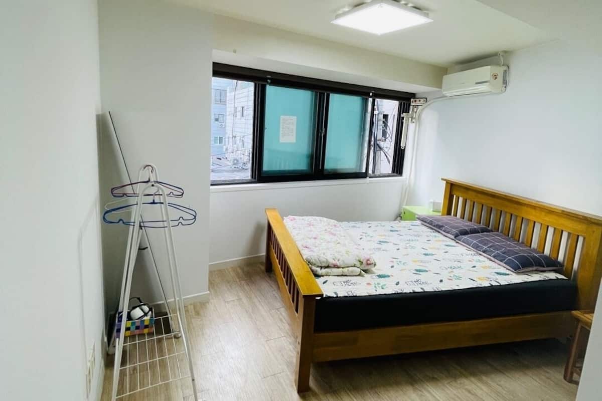 A bedroom with a wooden bed, a window with blinds, a radiator, and a clothes drying rack.