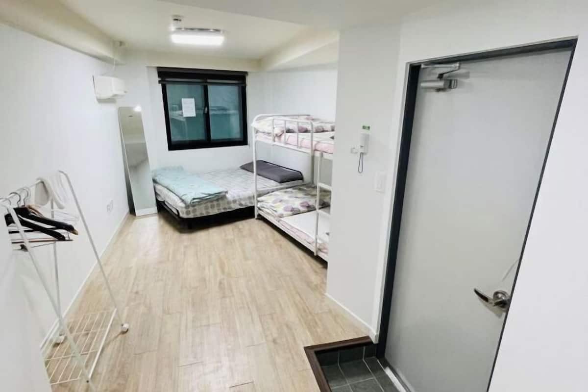 A small, single-room apartment with a bunk bed, a window, a door, and a laundry rack.