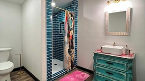 A bathroom with a white toilet, a teal vanity with a sink, a mirror with lights, a patterned rug, and a shower with blue tiles and a colorful shower curtain. - GUEST HOUSE "GROOVY" PRIVATE HOT TUB ON DECK, WIFI, ROKU. (Kennard)