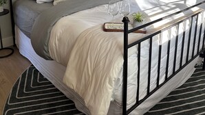 Een net opgemaakt bed met metalen frame, een nachtkastje met een lamp, en een raam met uitzicht op bomen.