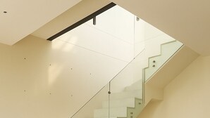 Un intérieur moderne avec un escalier en verre, un canapé beige et un petit coussin rose.