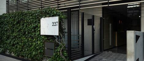 Entrada de un edificio moderno con un cartel con el número 227, una puerta metálica y un seto.