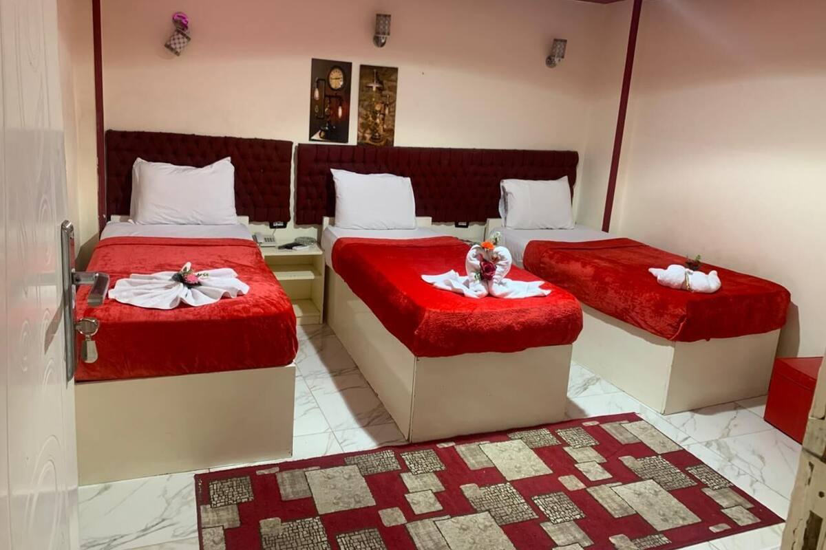 A hotel room with three beds, each with a red bedspread and white pillows, and a red and beige patterned rug on the floor.