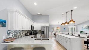 A spacious kitchen with white cabinets, a large island with bar stools, stainless steel appliances, and pendant lighting. - Tranquility where relaxation meets coastal charm (Marco Island)