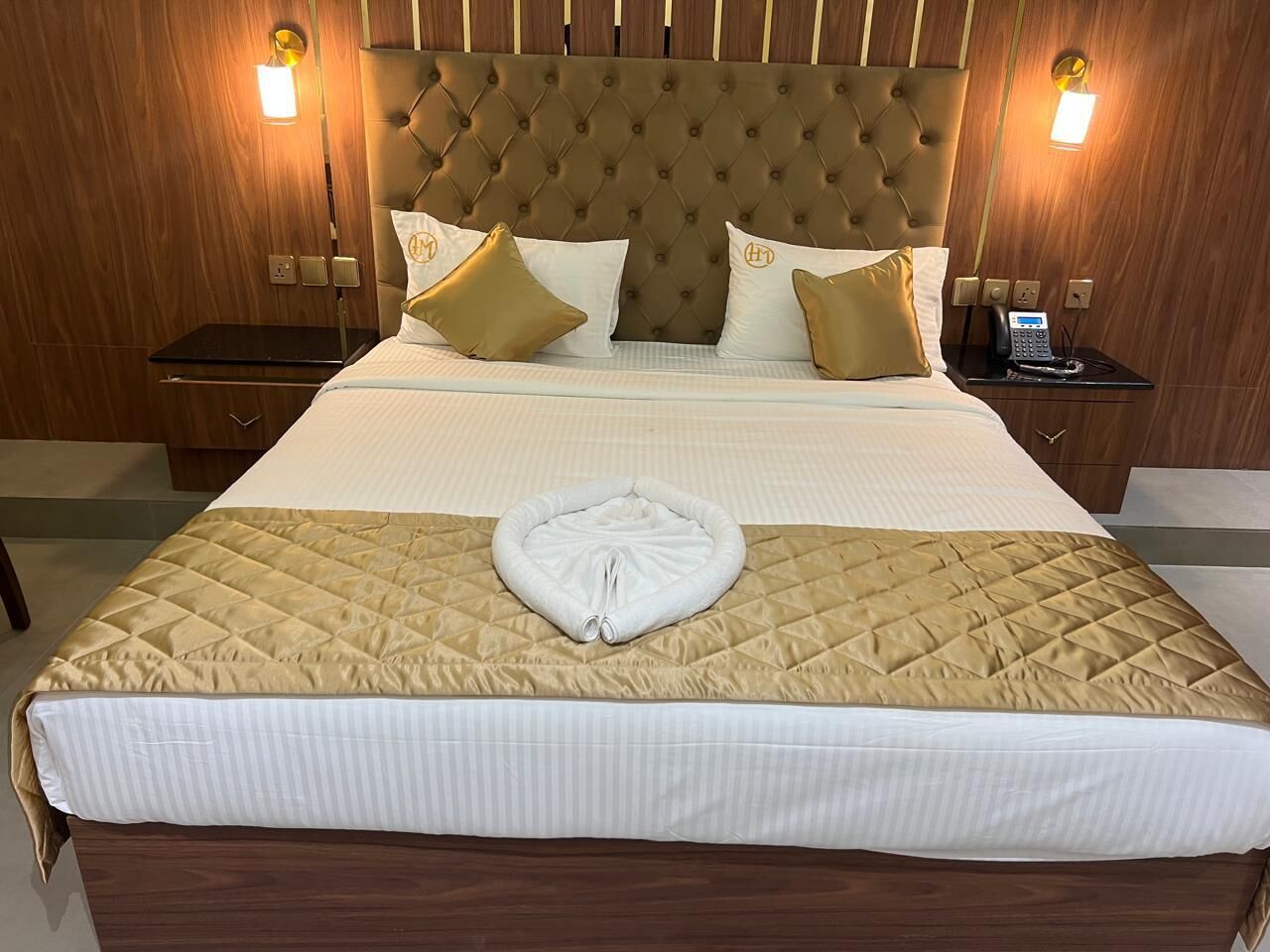 A neatly made bed with a quilted headboard, two bedside tables with lamps, and a visible power outlet on the wall.