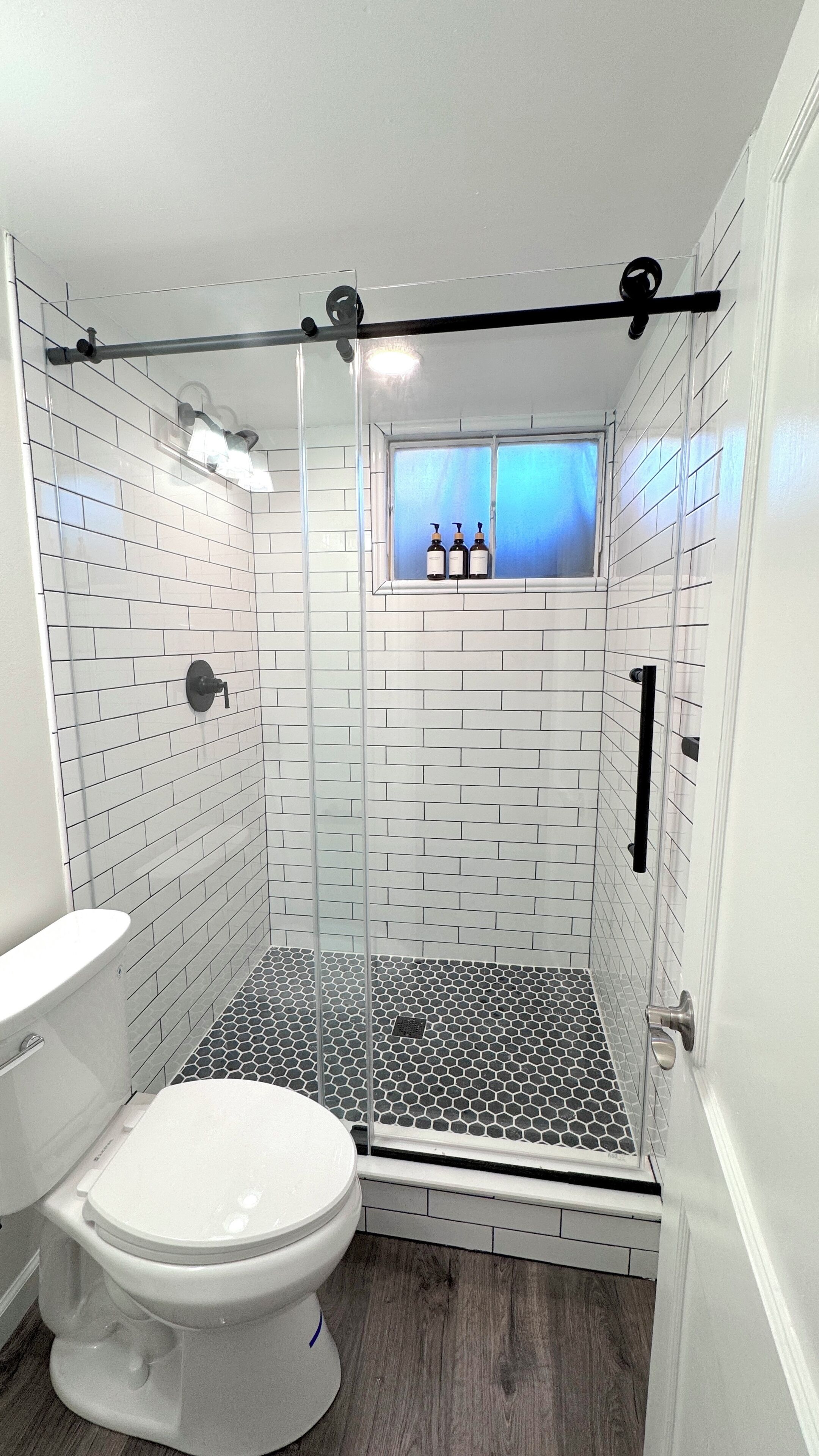 A bathroom with white subway tile walls, a black and white hexagonal floor, a glass shower enclosure, and a window with bottles on the sill.