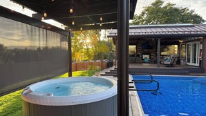 A hot tub and a swimming pool under a covered patio area with a view of a backyard and a wooden fence. - Fun House with Pool, Hot Tub, Cool Outdoor Living Space (Centennial)