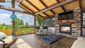An outdoor deck with wooden flooring, a stone fireplace, and a TV mounted on the wall. - Fun House with Pool, Hot Tub, Cool Outdoor Living Space (Centennial)