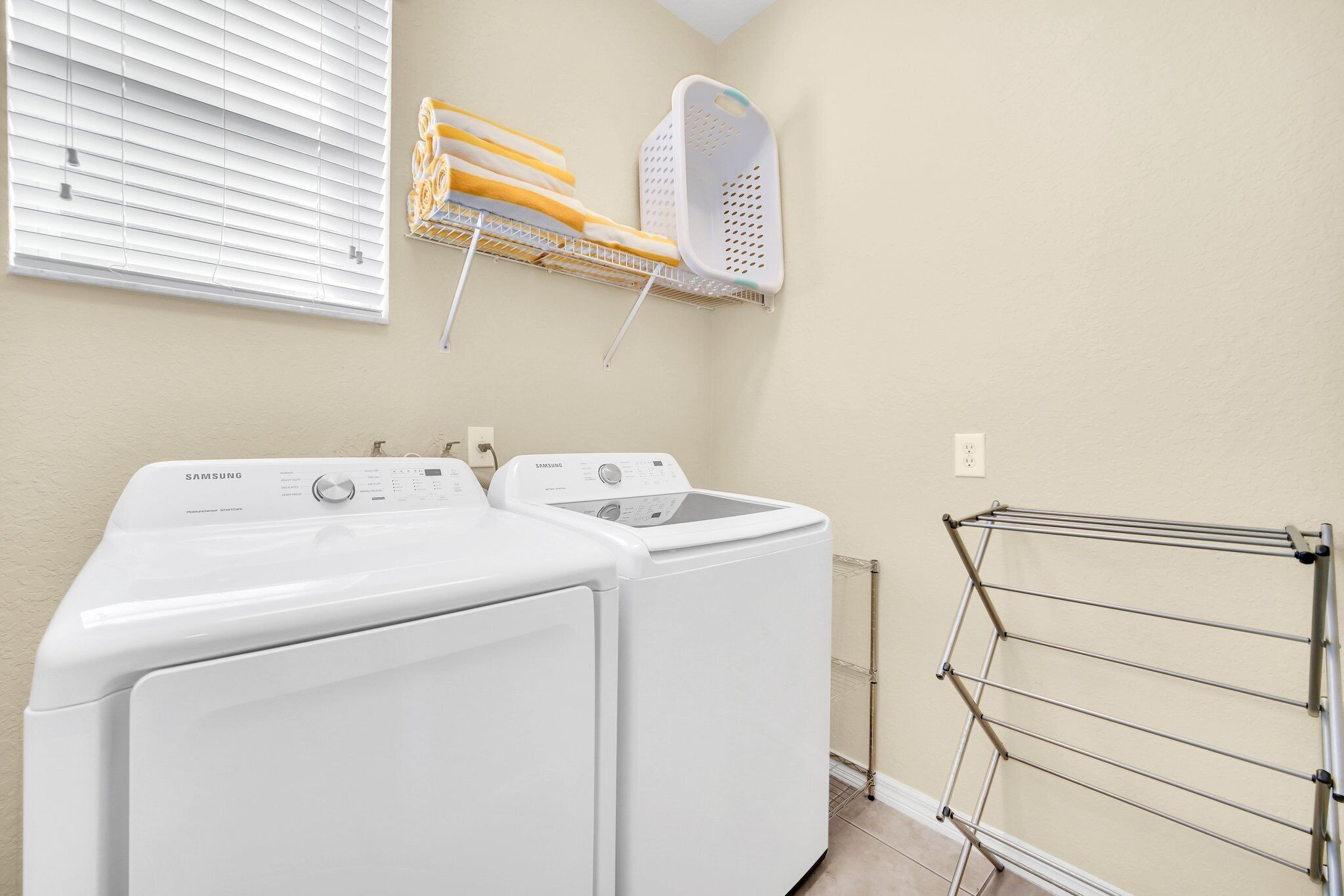 A laundry area with two white Samsung washing machines, a drying rack, and a wall-mounted laundry basket.