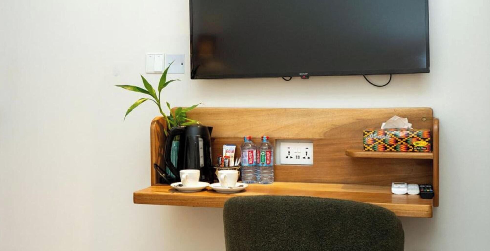 Ein Hotelzimmer mit einem Holzkonsolentisch, einem an der Wand befestigten Fernseher, einer Topfpflanze, einer Kaffeemaschine, Tassen, Wasserflaschen, einer Taschentuchbox und einer Fernbedienung.