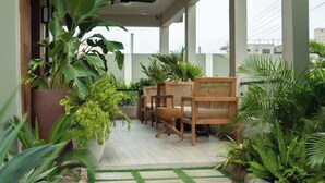 A covered outdoor seating area with wooden chairs and large potted plants.
