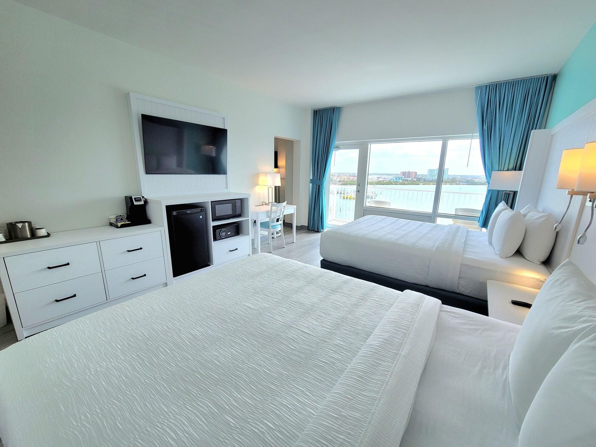 Habitación de hotel con cama, escritorio con televisor, mini refrigerador, lámpara y vista al agua.