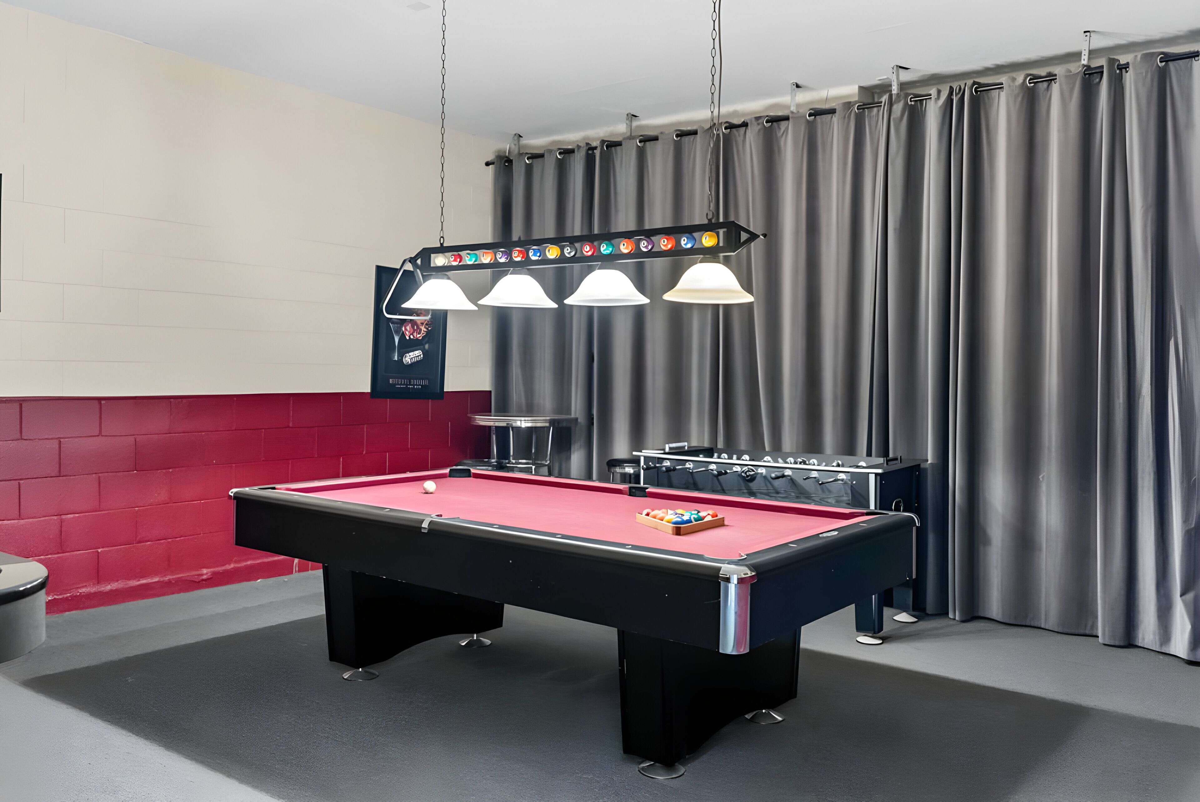 A pool table with a red felt surface, a foosball table, and a set of overhead lights.
