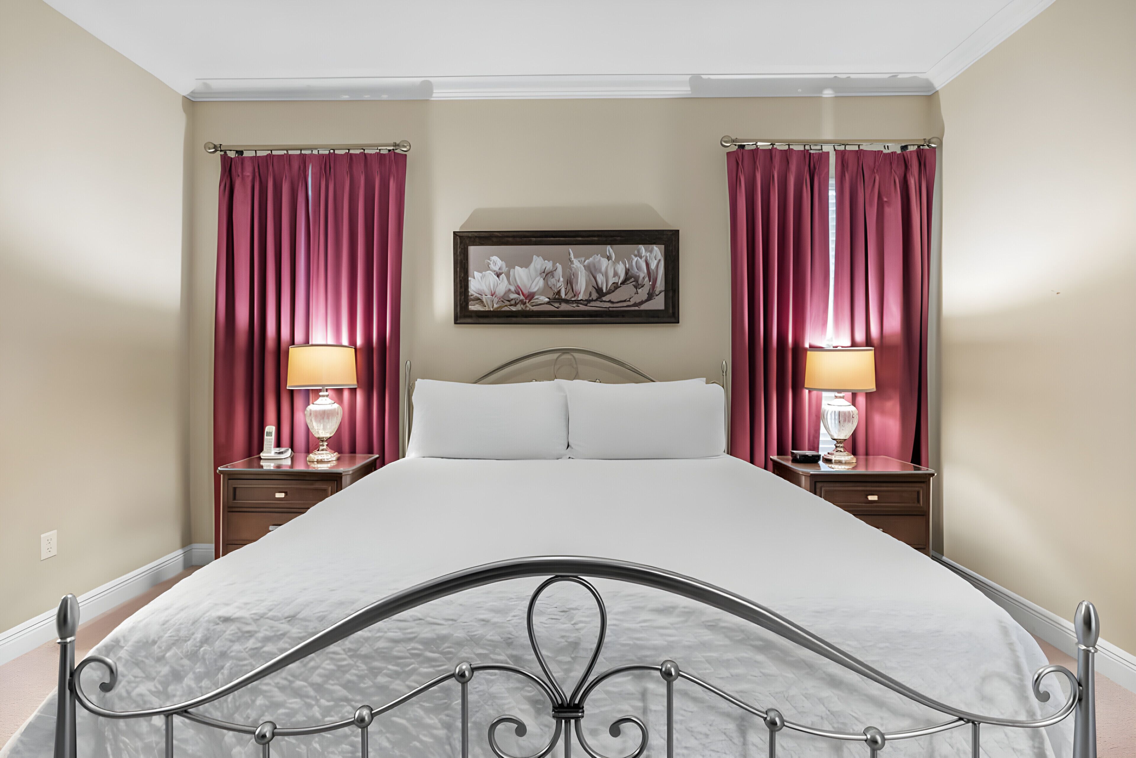 A neatly made bed with a metal frame, two bedside tables with lamps, and a framed floral artwork on the wall.
