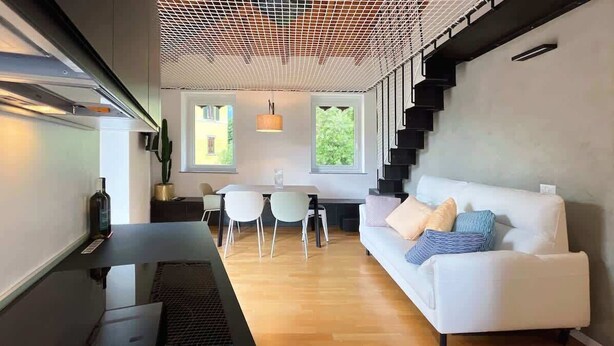 A modern living room with a white sofa, a dining table with chairs, a kitchen area, and a staircase. - Casa Iride - Affitti Brevi Italia (Lecco)