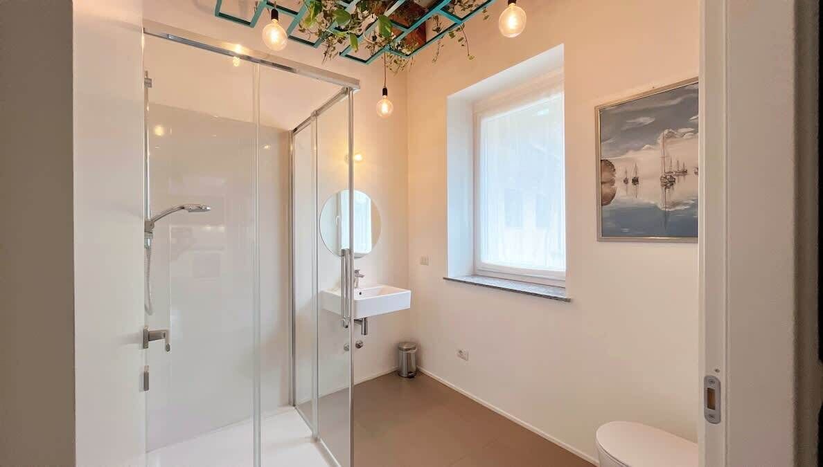 A modern bathroom with a glass shower enclosure, a sink, and a toilet. There is a window providing natural light and a framed artwork on the wall.