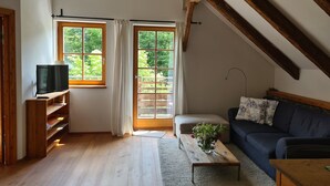 A cozy living room with a TV on a wooden stand, a sofa, a coffee table with flowers, and large windows with curtains.