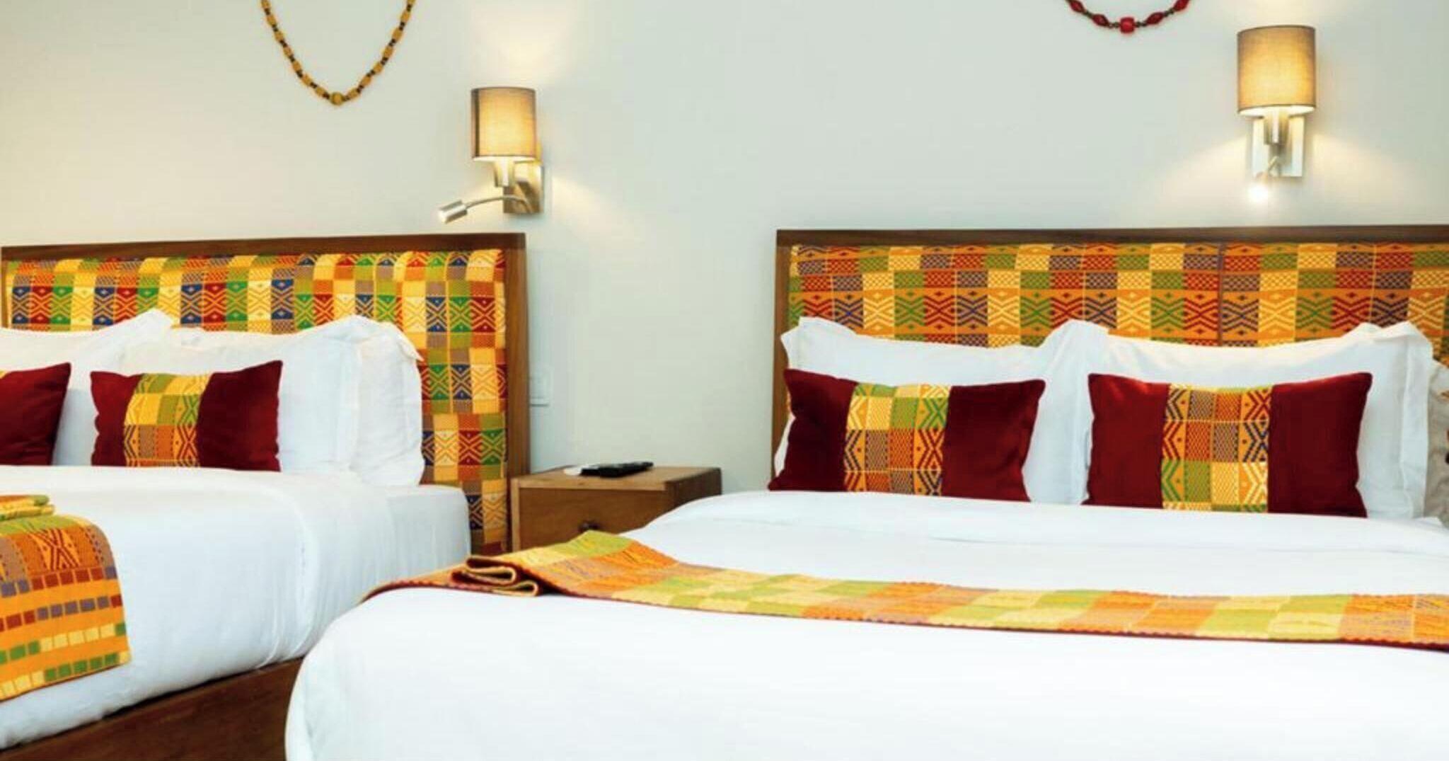 Habitación de hotel con dos camas, cada una con un cabecero estampado, ropa de cama blanca y almohadas rojas. Hay dos lámparas de pared y una mesita de noche pequeña con un control remoto.