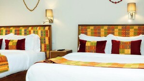 A hotel room with two beds, each with a patterned headboard, white bedding, and red pillows. There are two wall-mounted lamps and a small bedside table with a remote control.