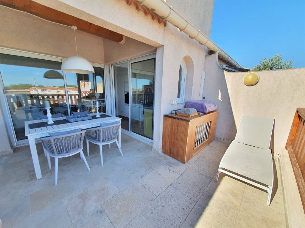 Une terrasse extérieure avec une table et des chaises, un banc en bois et une porte vitrée donnant sur une pièce.