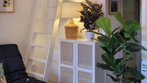 Ein gemütliches Wohnzimmer mit einer schwarzen Couch, einem gelben Tisch mit Flasche und Glas, einer weißen Kommode mit Pflanzen, einer Leiter zu einem Schlafplatz im oberen Bereich und einer Tür zu einem anderen Raum.