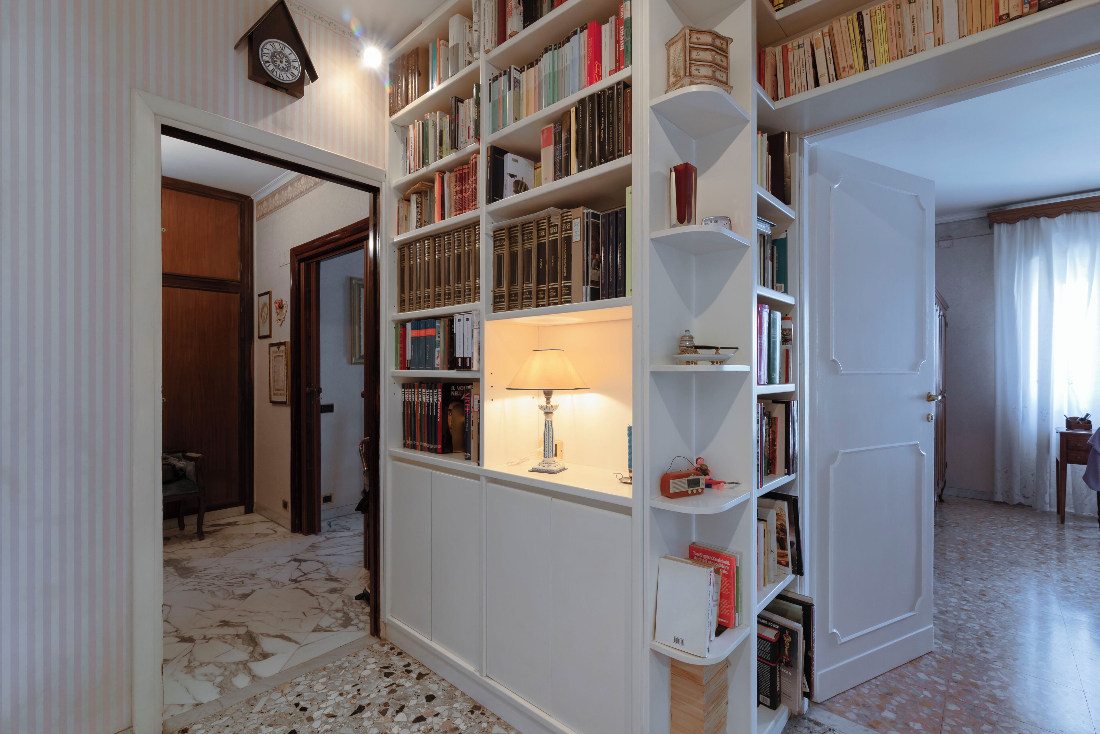 A room with a marble floor, a bookshelf filled with books, a lamp, and a clock on the wall.