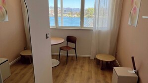 A modern room with a large mirror, a wooden floor, a round table, a chair, and a view of a body of water through large windows. - Comfort Appt in a Premium Residence- Sport/cinema (Huningue)
