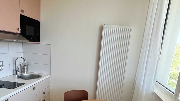 A small, modern kitchen with white cabinets, a microwave, and a sink. There is a round wooden table with two chairs and a radiator next to a window. - Comfort Appt in a Premium Residence- Sport/cinema (Huningue)