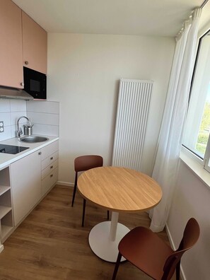 A small, modern kitchen with white cabinets, a microwave, and a sink. There is a round wooden table with two chairs and a radiator next to a window. - Comfort Appt in a Premium Residence- Sport/cinema (Huningue)