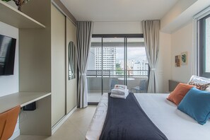 A hotel room with a bed, a TV, a balcony with a view of buildings, and a chair.