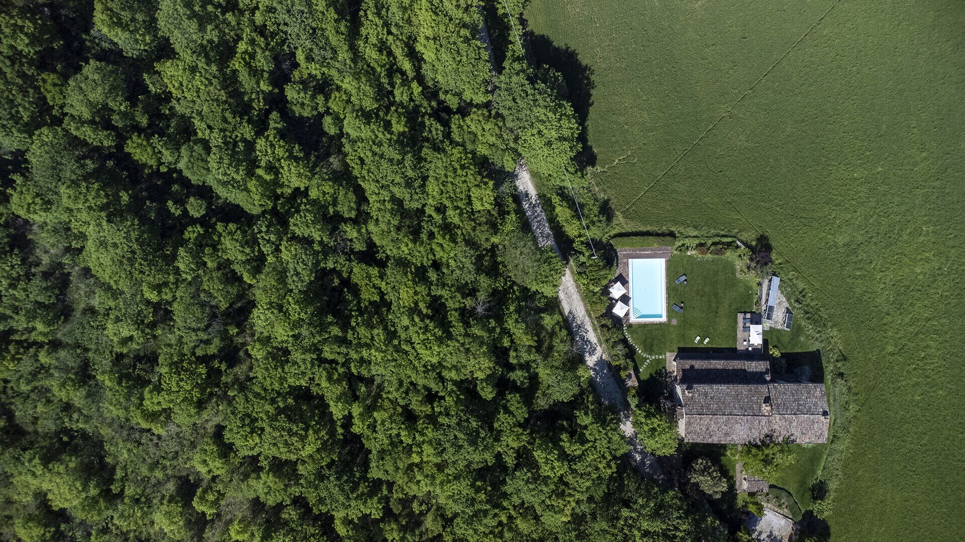 An aerial view of a property with a swimming pool, a small house, and dense forest surrounding it.