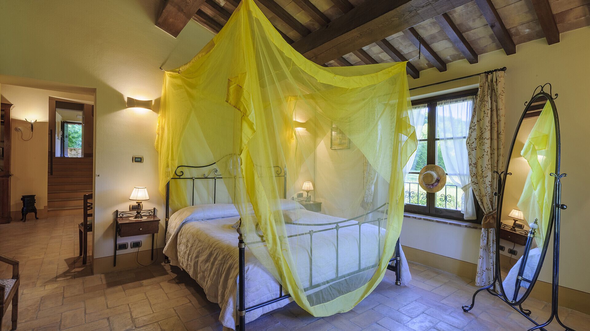 Een slaapkamer met een bed onder een geel hemelbed, een nachtkastje met een lamp, een raam met gordijnen en een spiegel op een standaard.