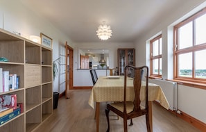 A dining area with a table set for four, a bookshelf, and a view through the window. - Pipeland Farm Steading (No 4) (St Andrews)