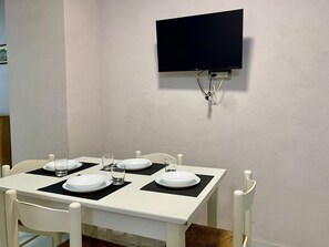 A dining area with a white table set for four, a flat-screen TV mounted on the wall, and a white chair with a woven seat cushion. - House Of Fabio | A traditional townhouse in the historic center, surrounded by art and history (Napoli)