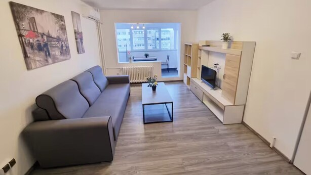 Una sala de estar moderna con un sofá gris, una mesa de centro y un televisor sobre un mueble de madera. Hay dos cuadros en la pared y una ventana con vistas a edificios.