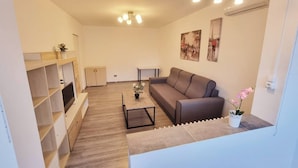 Una sala de estar moderna con sofá, una mesa de centro y un televisor sobre un mueble de madera. Hay luces fijadas en la pared y un ventilador de techo.