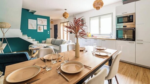 Una cocina moderna con armarios blancos, una mesa de comedor de madera dispuesta con platos y vasos, y una escalera que lleva a una sala de estar color turquesa.