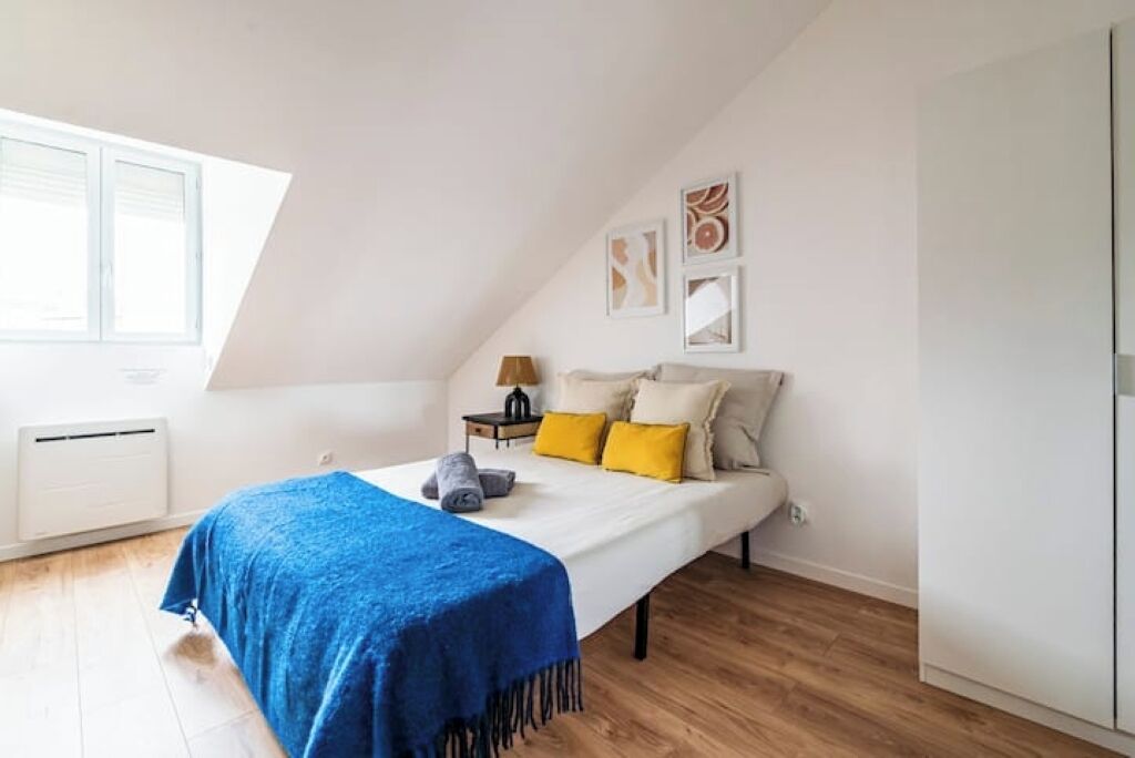 A bedroom with a sloped ceiling, a bed with a blue blanket, a nightstand with a lamp, and artwork on the wall.