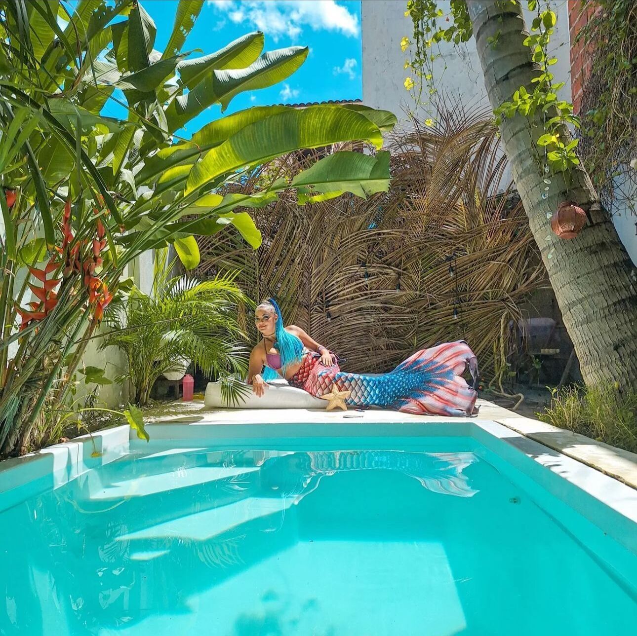 Une personne déguisée en sirène, allongée dans une piscine, entourée de plantes tropicales.