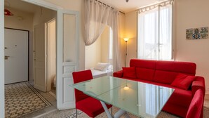 A room with a glass table, red chairs, a red sofa, a bed with white bedding, a floor lamp, and a door leading to another room. - La Terrazza di Apollo (Syracuse)