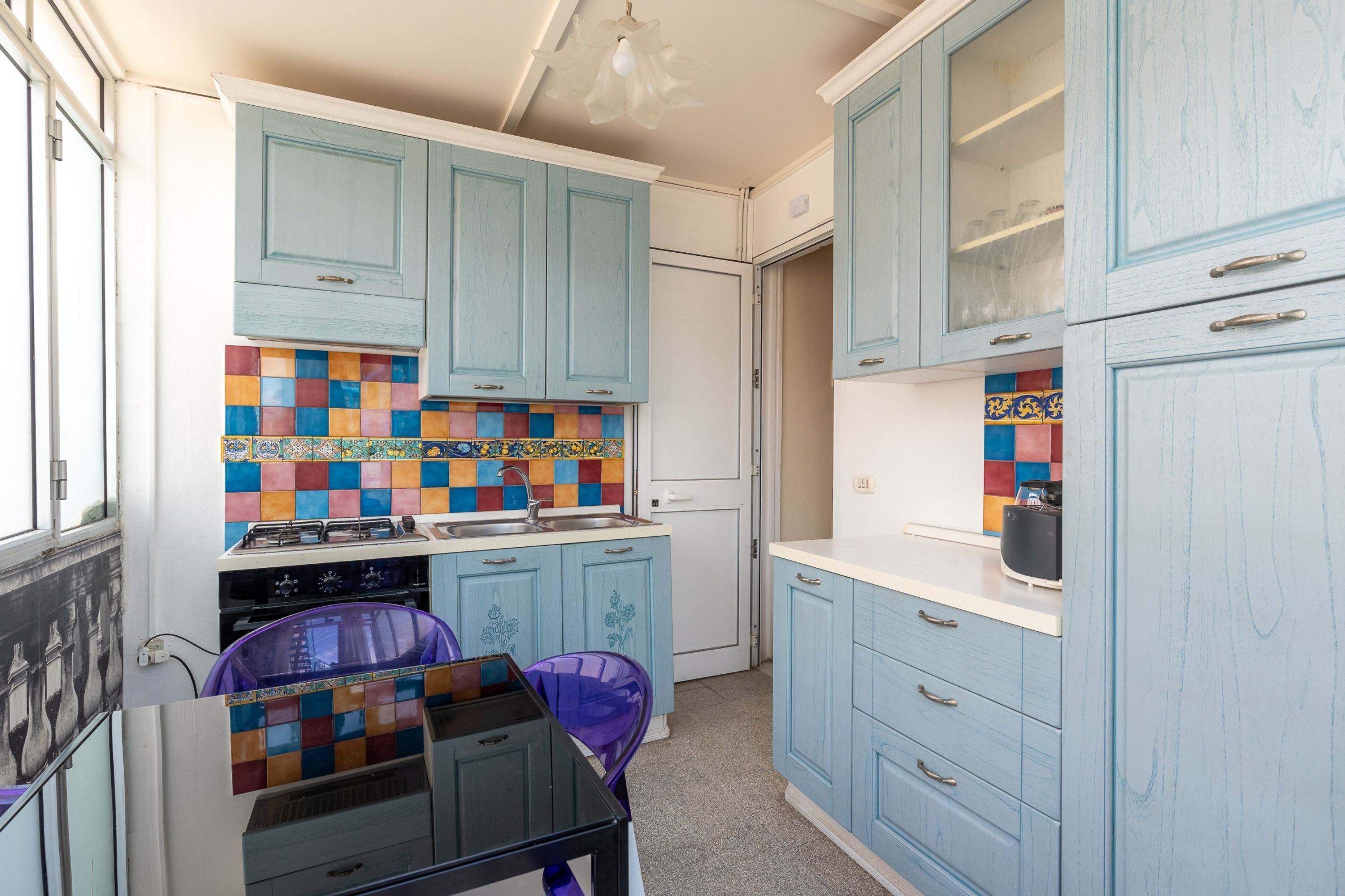 A compact kitchen with teal cabinets, a colorful backsplash, and a small dining area with a purple chair.